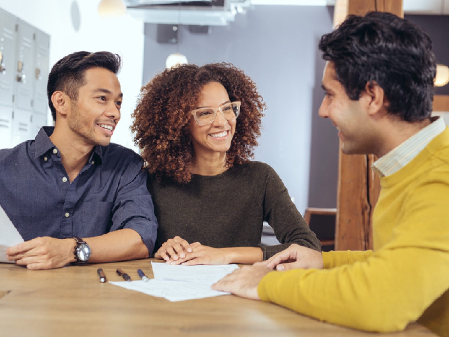 A couple is having a pleasant conversation with a person across the table, with papers in front of them, in what appears to be a meeting or consultation.