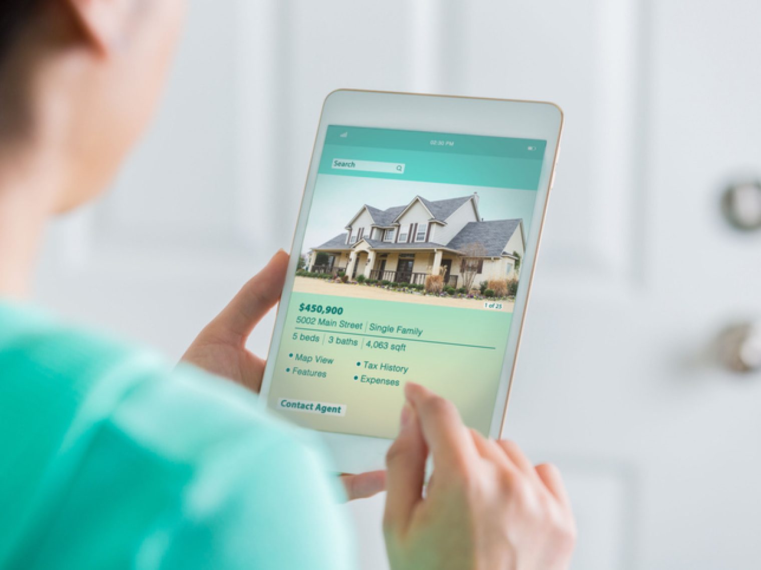 A person holds a tablet displaying a real estate app with a listing for a single-family home, suggesting they are searching for or interested in buying property.